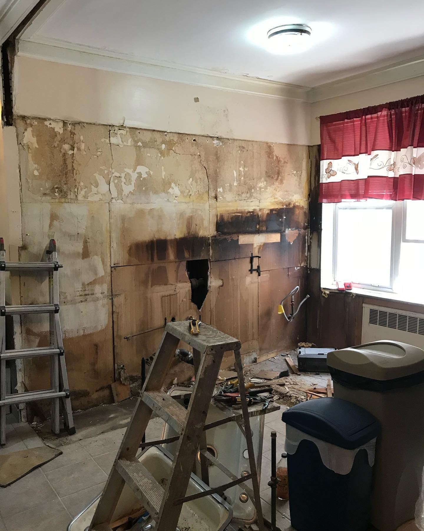 Kitchen renovation in progress: exposed walls, tools, ladder, trash cans. Window with red curtains on right.