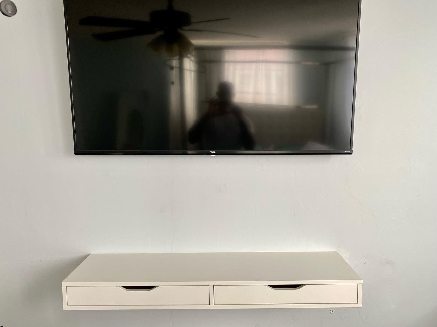 White wall-mounted TV and floating drawer unit below it. A person is reflected in the TV screen.