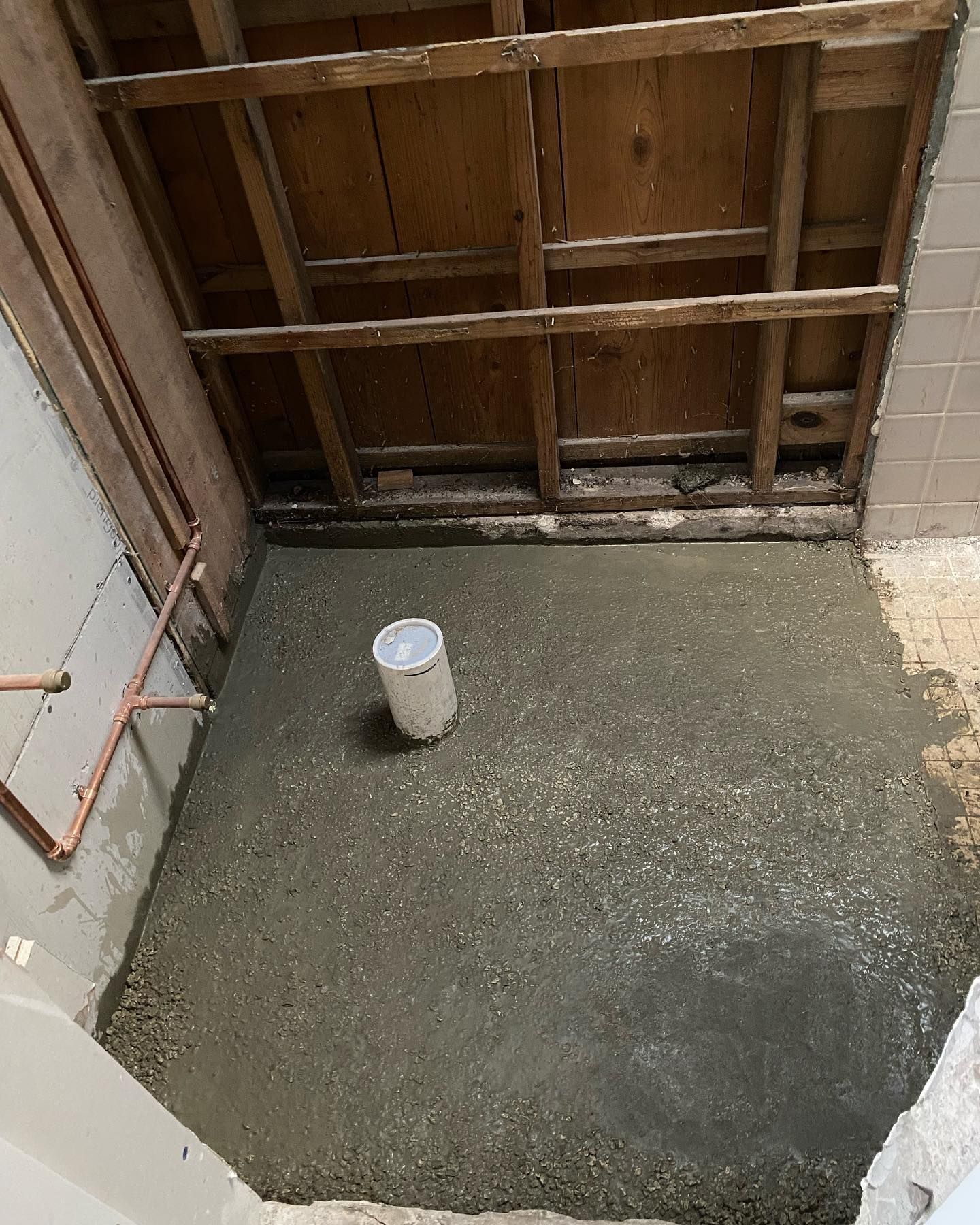 Bathroom renovation: unfinished floor with aggregate, exposed framing, and a bucket. Copper pipes and partially tiled walls.