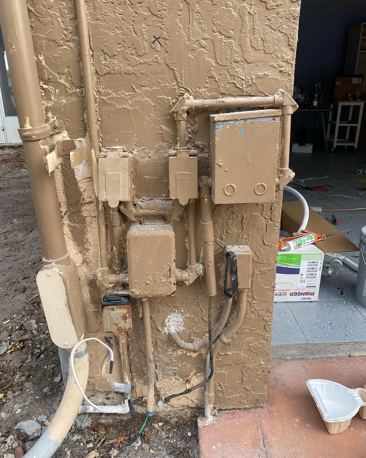 Brown painted electrical boxes and pipes on a stucco wall.