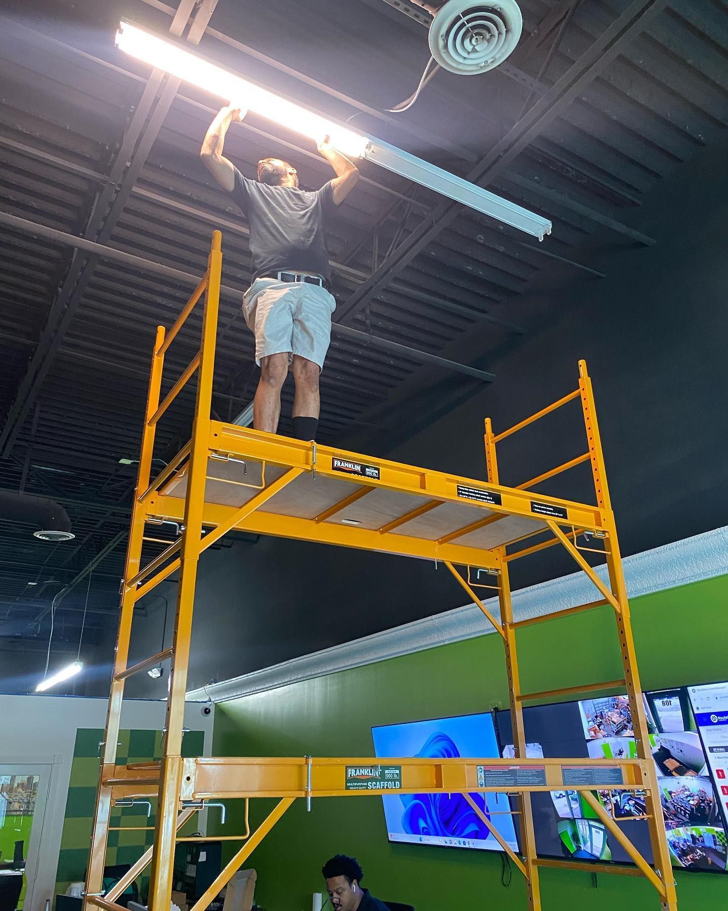 Person on a yellow scaffold installs a fluorescent light in a room with a dark ceiling and green walls.