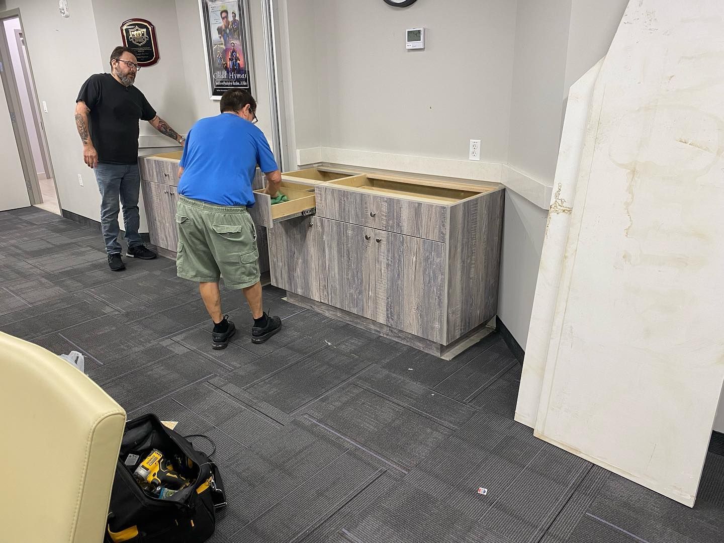 Two men installing a cabinet in a room with gray carpet; one man is holding a box.