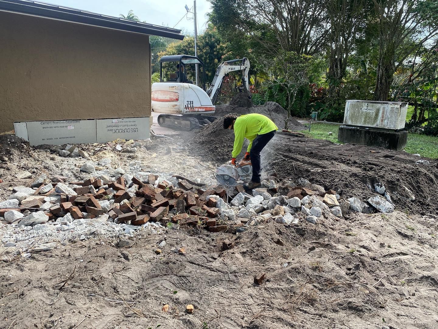 A worker in a yellow shirt breaks rocks with a pickaxe next to a small excavator and building.