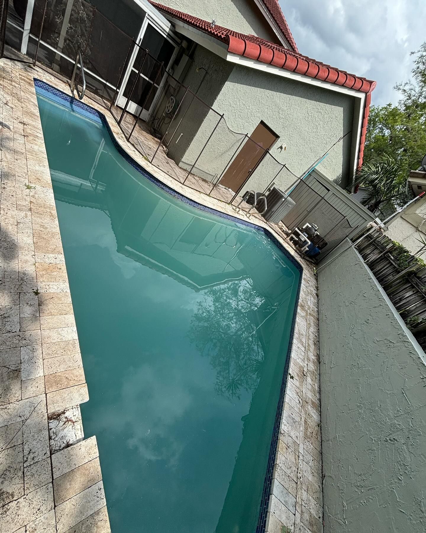 A teal-colored swimming pool surrounded by beige stone and a building with a red roof.