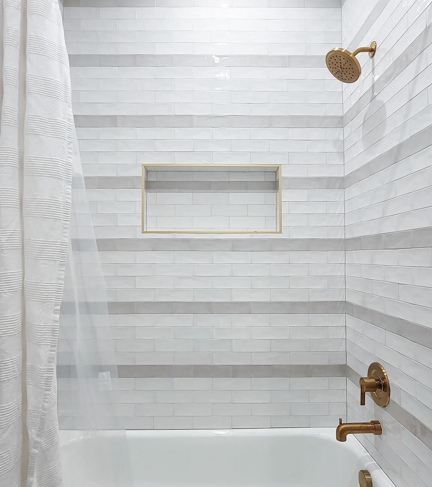 Shower with white and grey striped tile, gold fixtures, and white shower curtain.