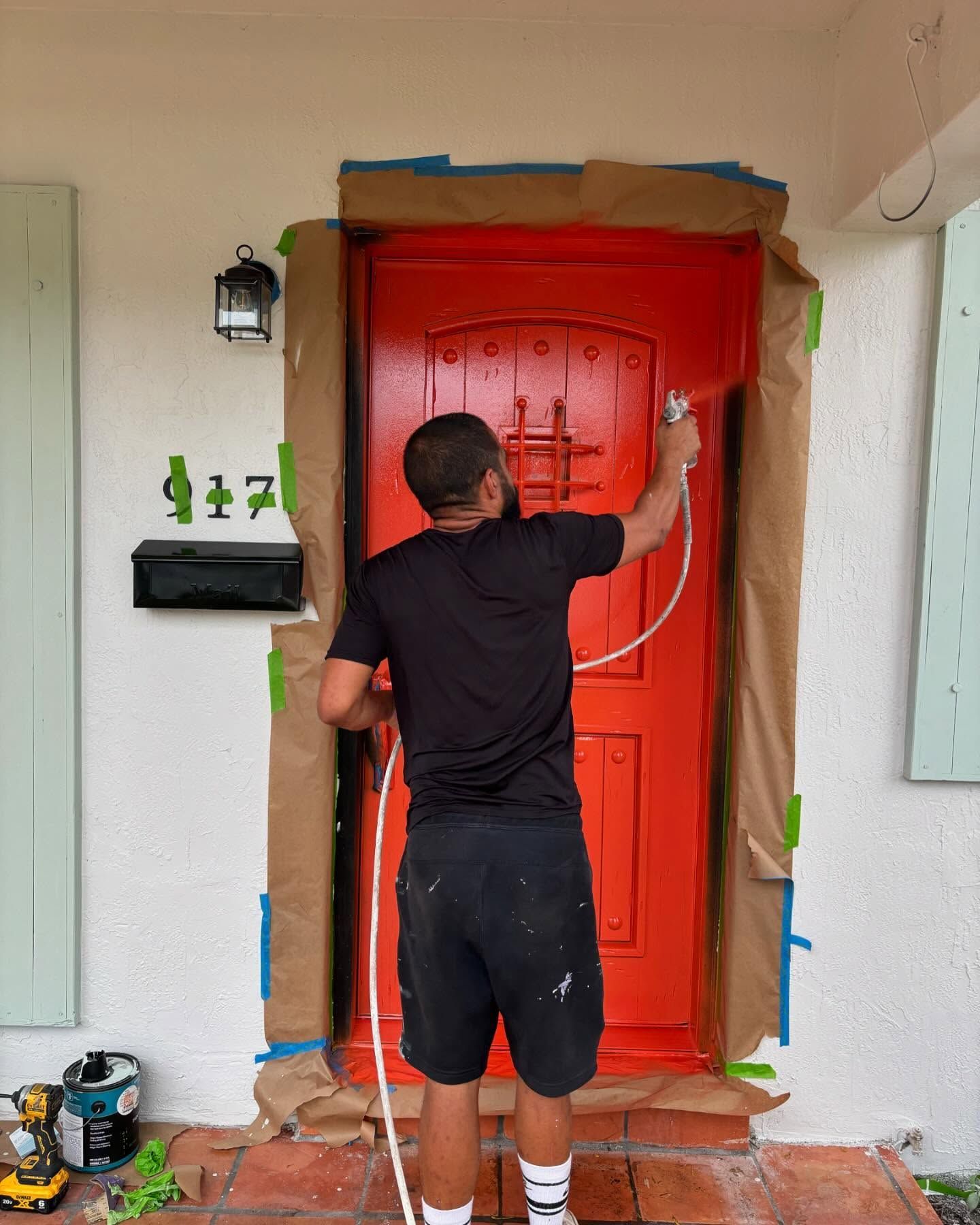 Man spraying bright orange paint on a front door, masked with brown paper. Outdoors.