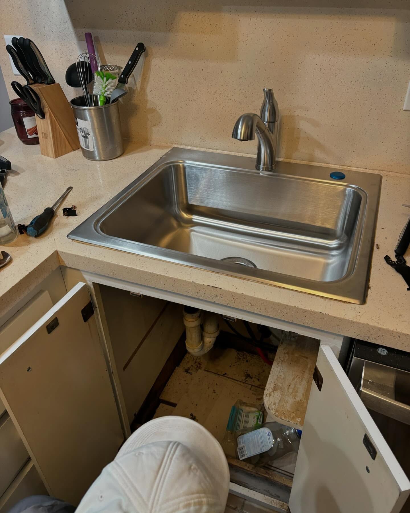 A small kitchen sink, installed in a countertop, with open cabinet doors and tools.