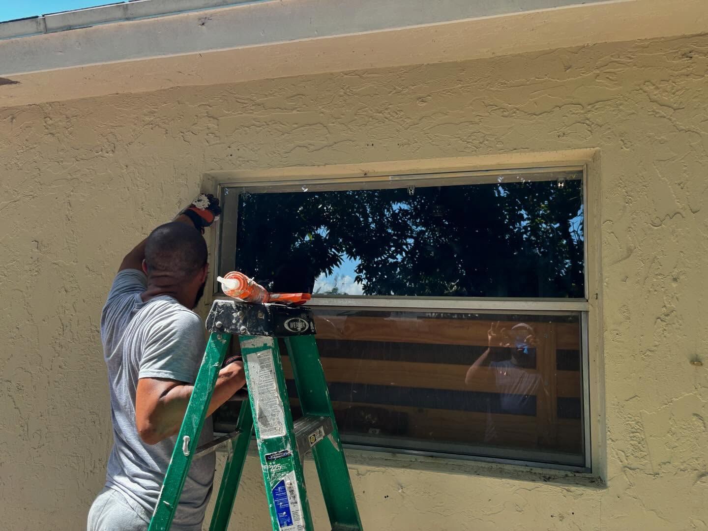 Man on a ladder caulking a window on a beige stucco building.