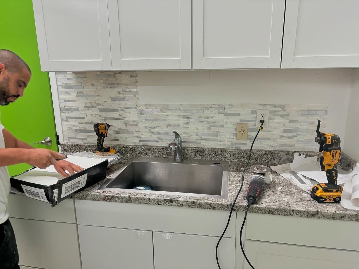 Man installing backsplash tiles in a kitchen, below white cabinets and above a sink.