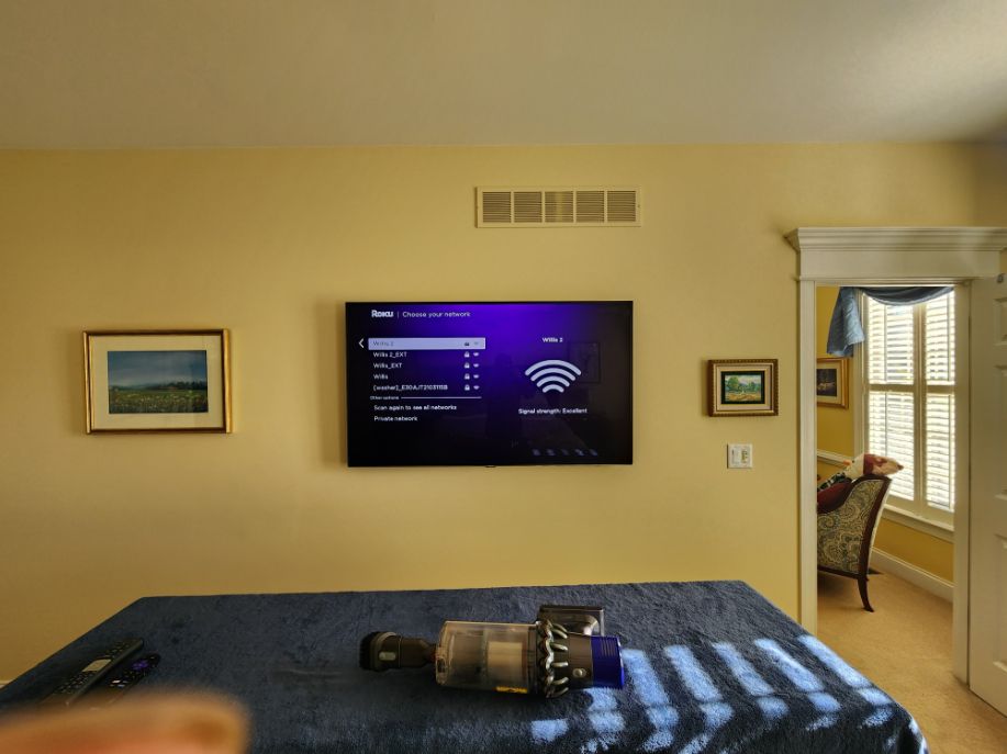 Bedroom with wall-mounted TV above bed, framed art, and a doorway to a sunlit room.
