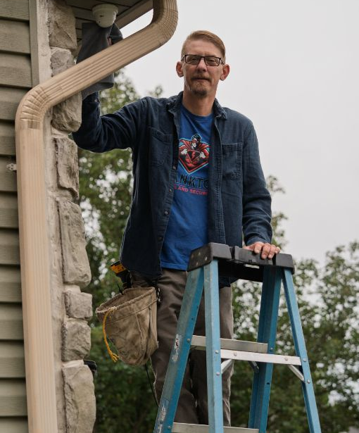 Man standing on a ladder repairing the exterior trim of a house, holding a tool