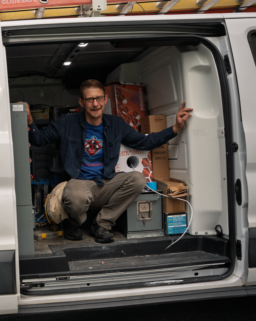 Man crouching in the back of a white cargo van, surrounded by tools and boxes, with the van doors open.