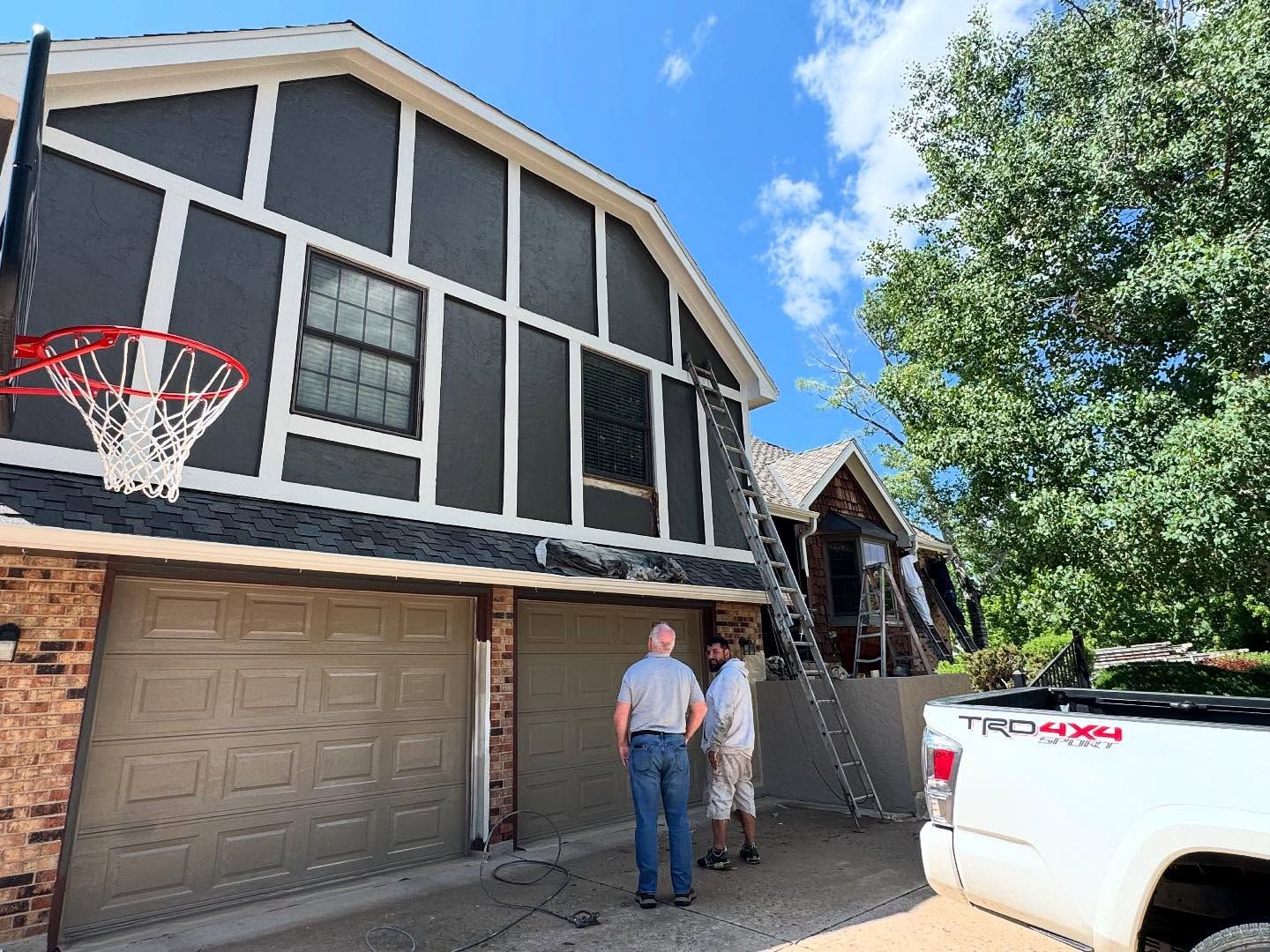 Two people standing by a garage and white truck outside a two-story house with a basketball hoop