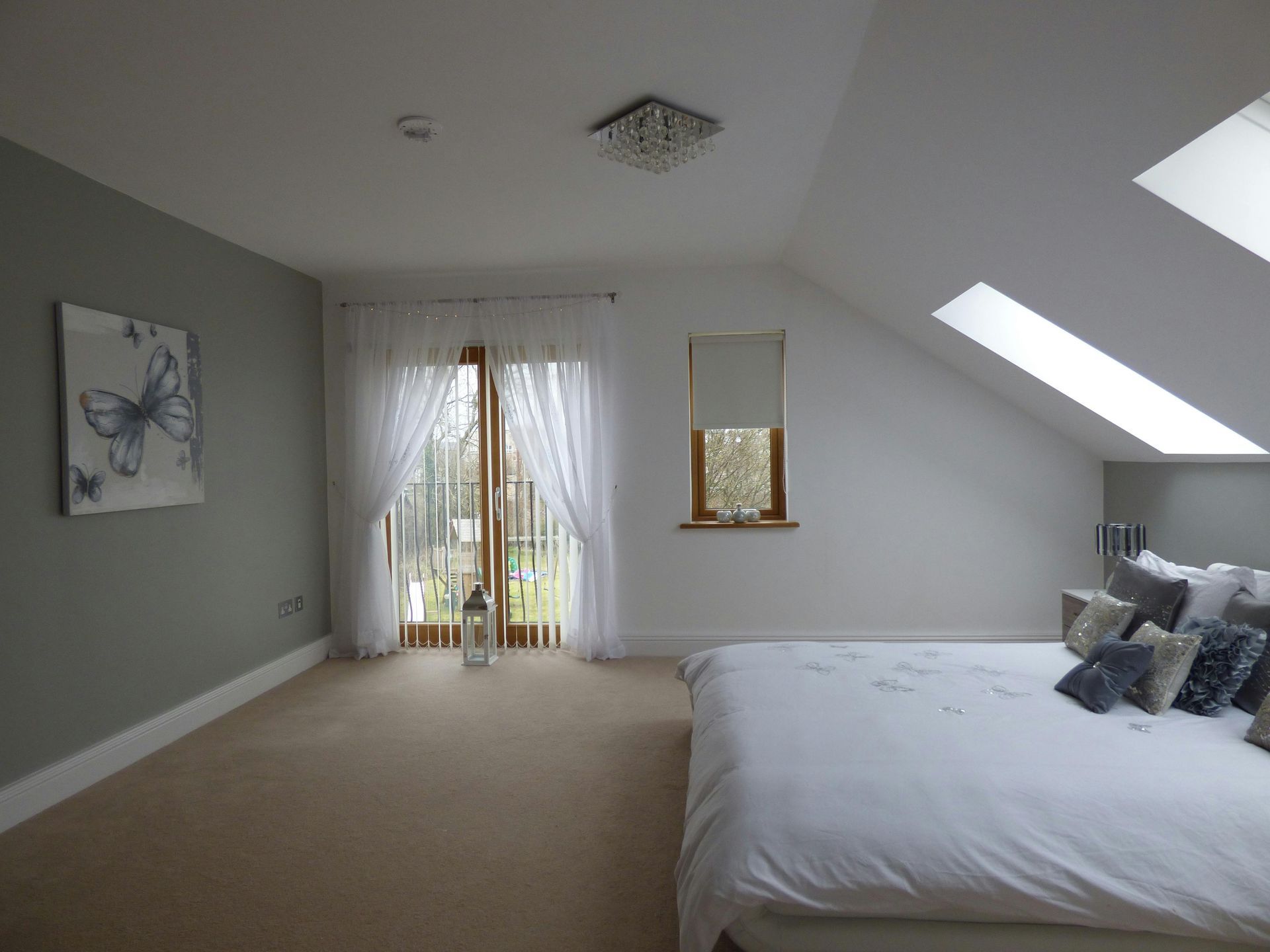 Bright attic bedroom with a skylight, white bed, beige carpet, and sheer curtains over balcony doors