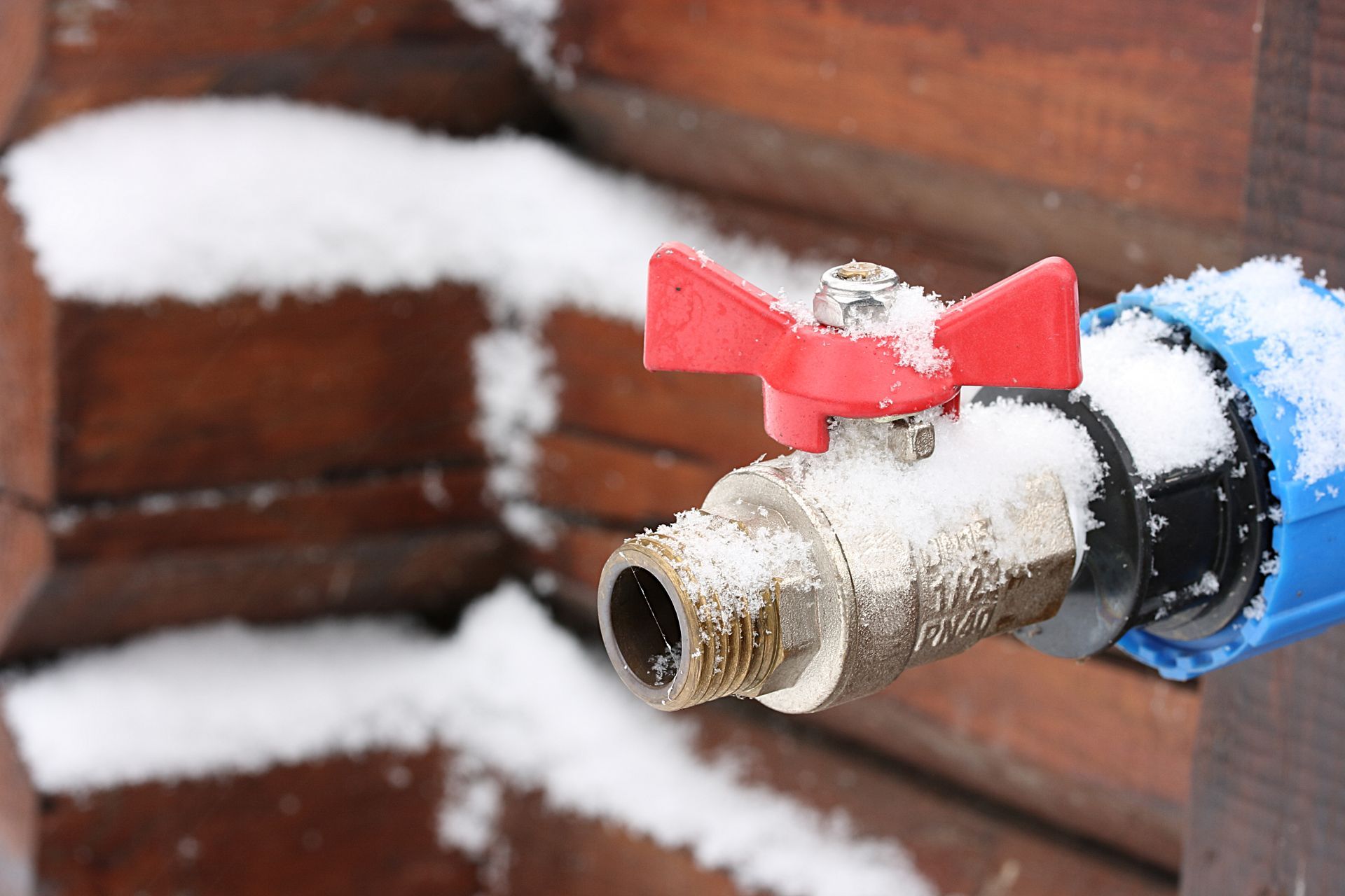 A snow-covered blue and red water valve mounted on a wooden fence outdoors on a winter day.