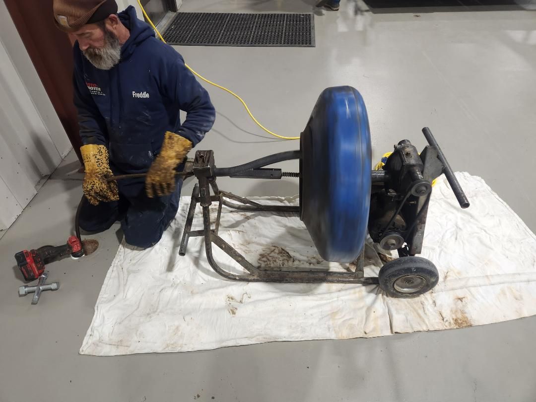 Man kneels beside a blue wheel and tool-filled machine on a white tarp in a workshop.