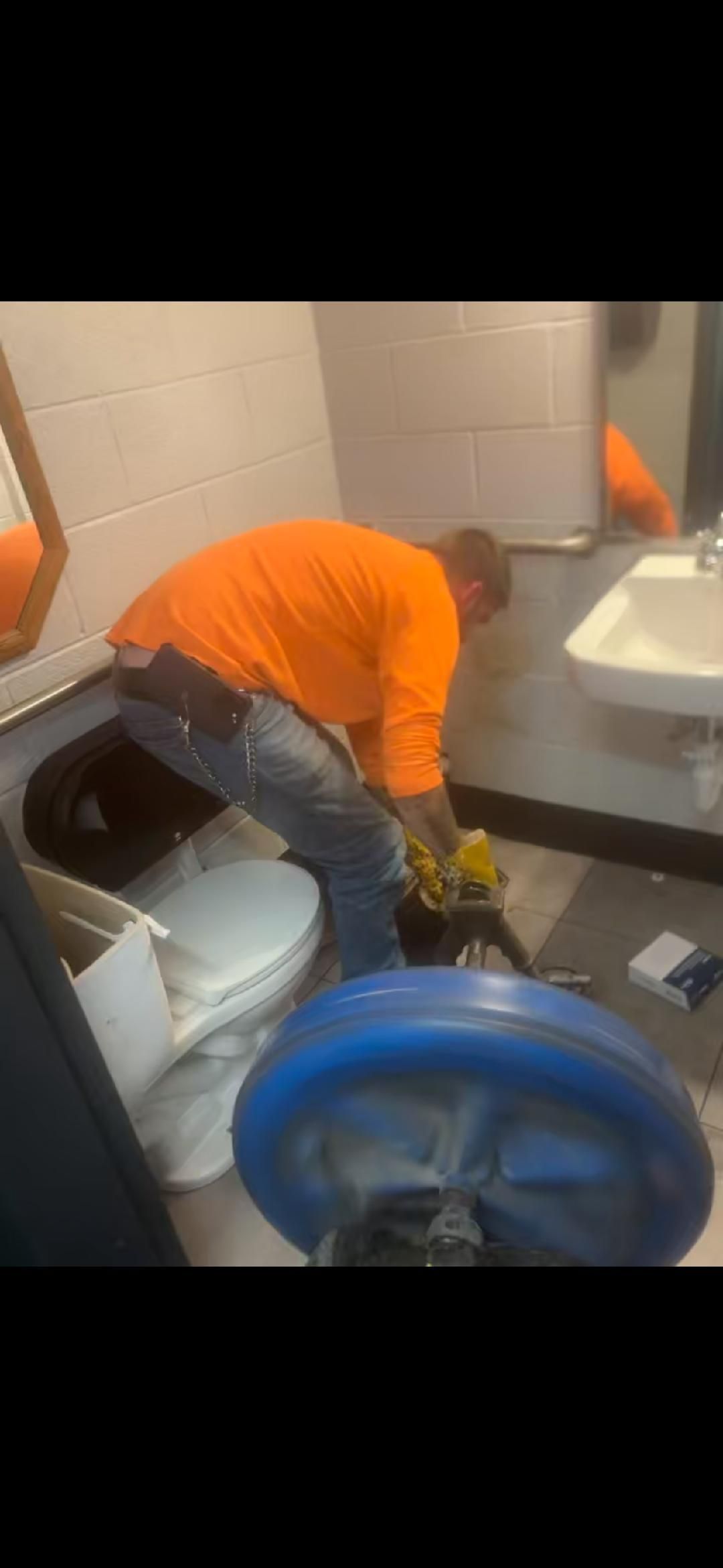 Person in orange shirt crouches in a bathroom, cleaning near a toilet and mop bucket.