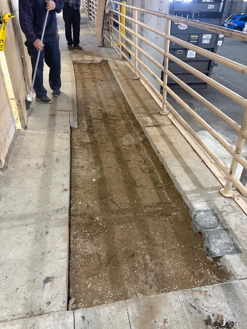 Narrow sidewalk with a muddy construction trench beside metal railings; two people stand at the left.