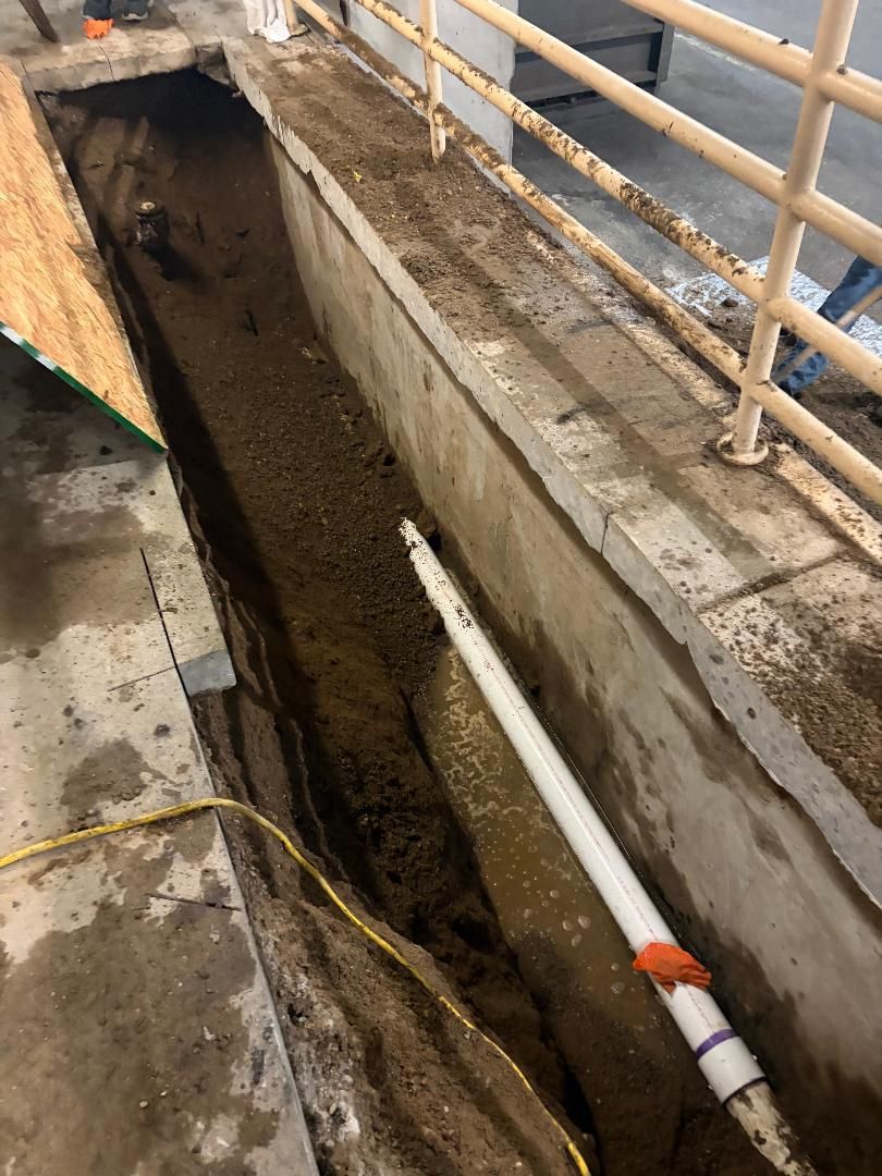 Narrow construction trench beside a concrete walkway, with metal railings and exposed pipes.