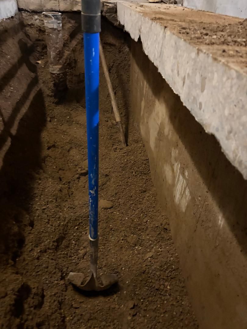 Blue-handled shovel standing in a narrow dirt trench beside a concrete wall.