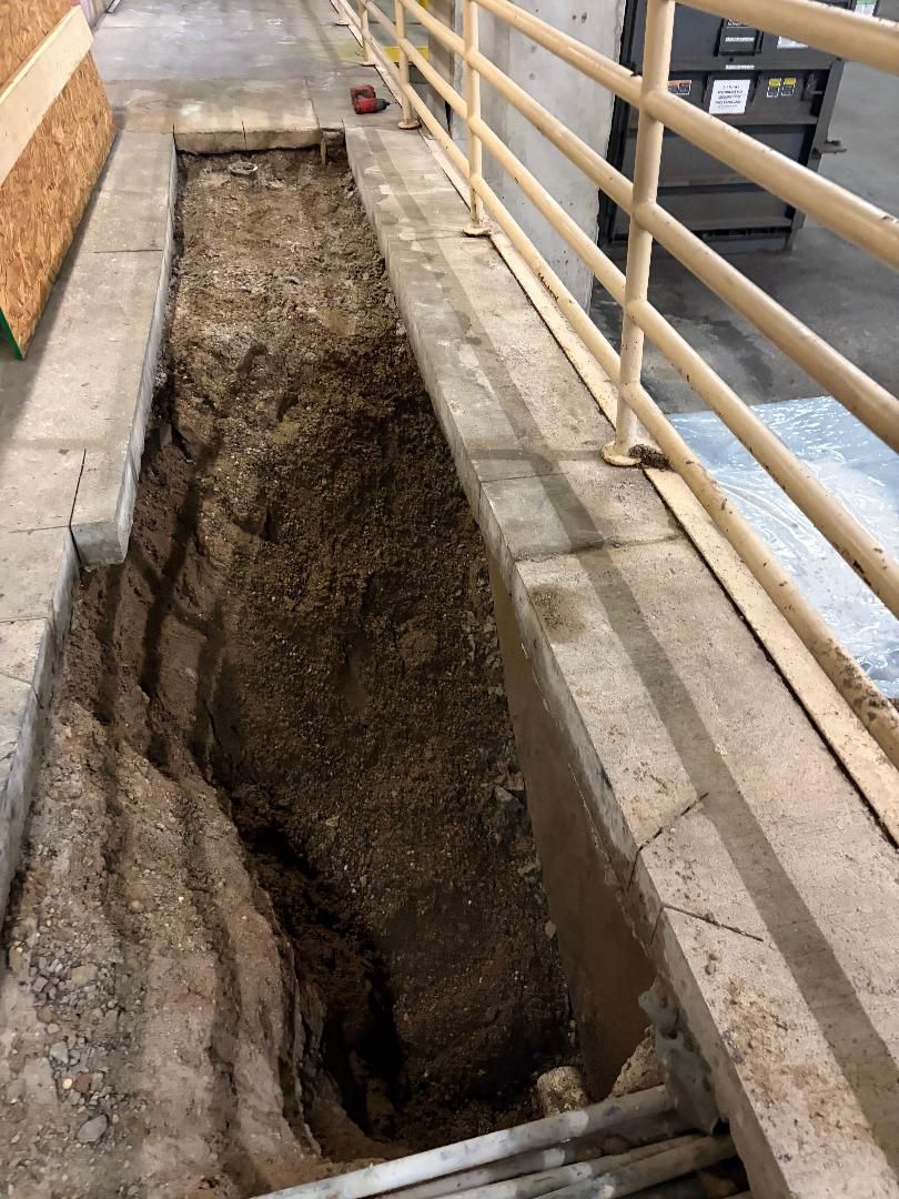 Deep trench cut into a concrete walkway beside a metal railing and brick wall.