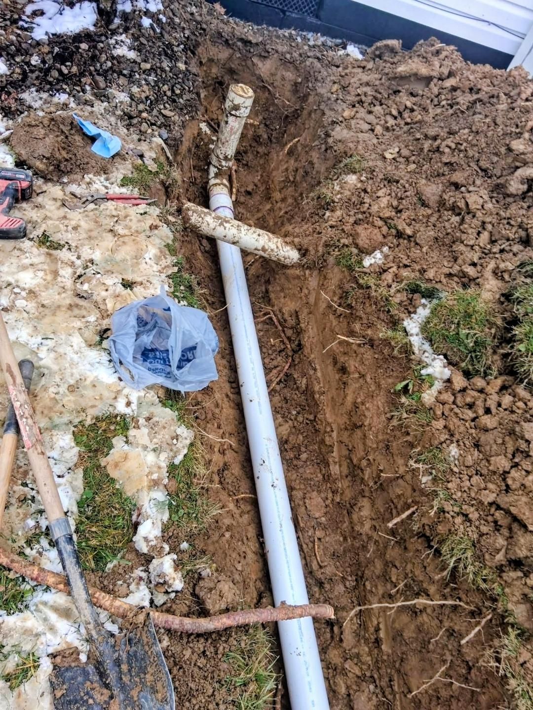 Excavation trench with exposed white pipe, muddy soil, and a plastic bag in debris-covered ground.