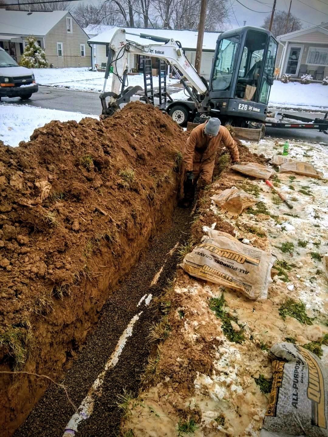 Backhoe excavating a long trench beside a snowy residential street, with houses in the background