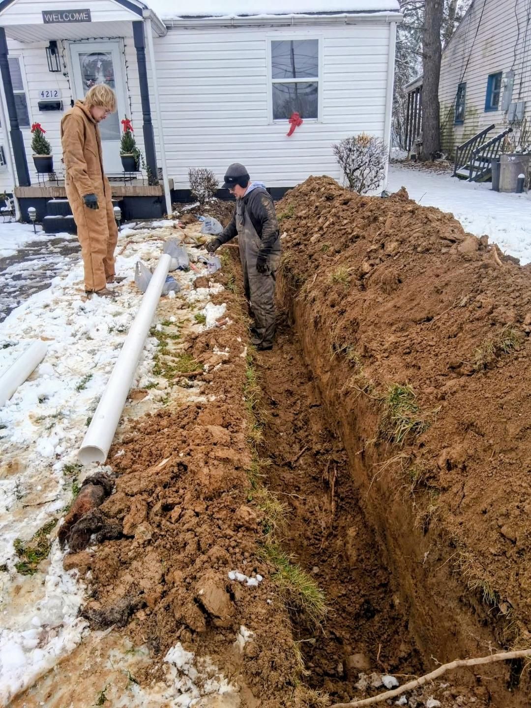 Workers dig a deep trench beside a white house in a snowy yard