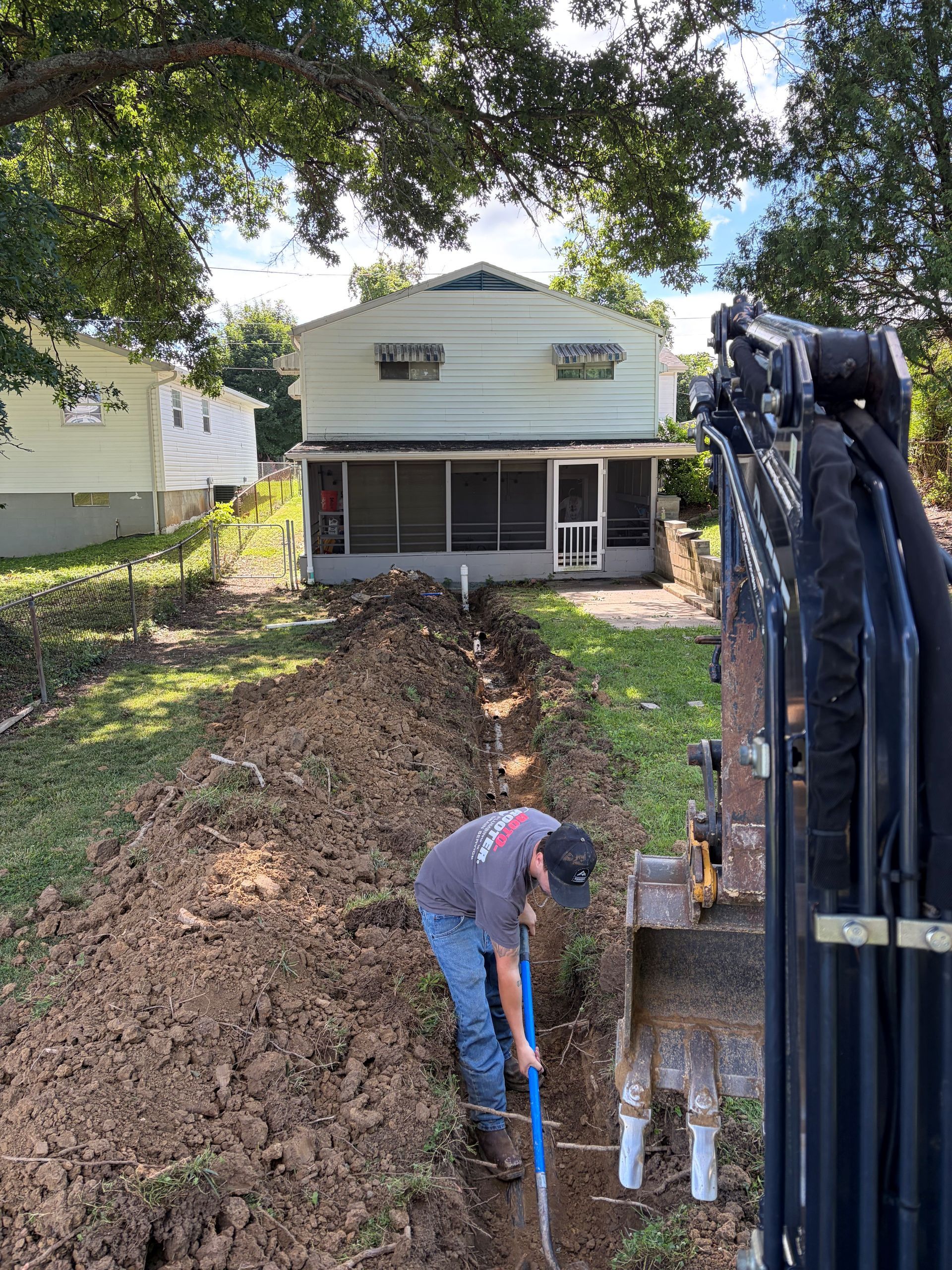 Worker in blue shirt shovels dirt beside a house during excavation, with an excavator on the right.