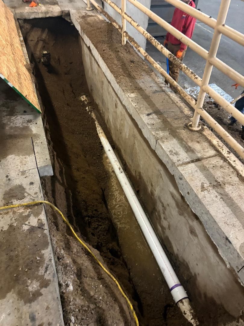 Open trench beside a concrete walkway with a white pipe and safety railings above