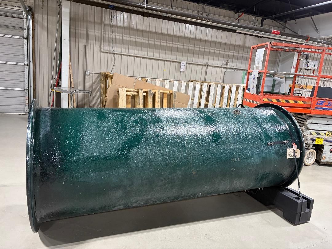 Large dark green cylindrical tank in a warehouse, resting on a black stand with a lift in the background
