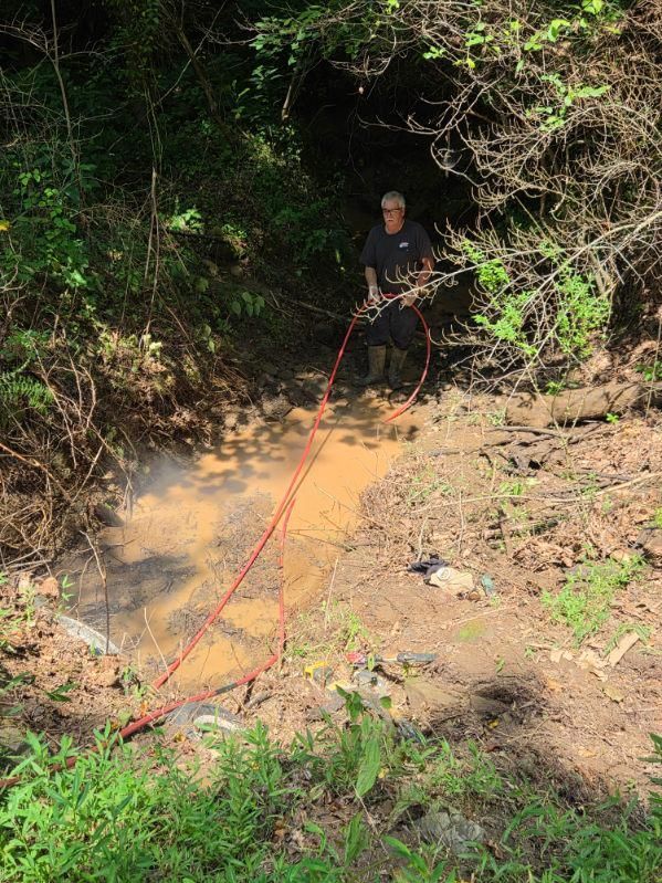 Person standing in a muddy ravine, handling a red hose amid dust or smoke and dense greenery.
