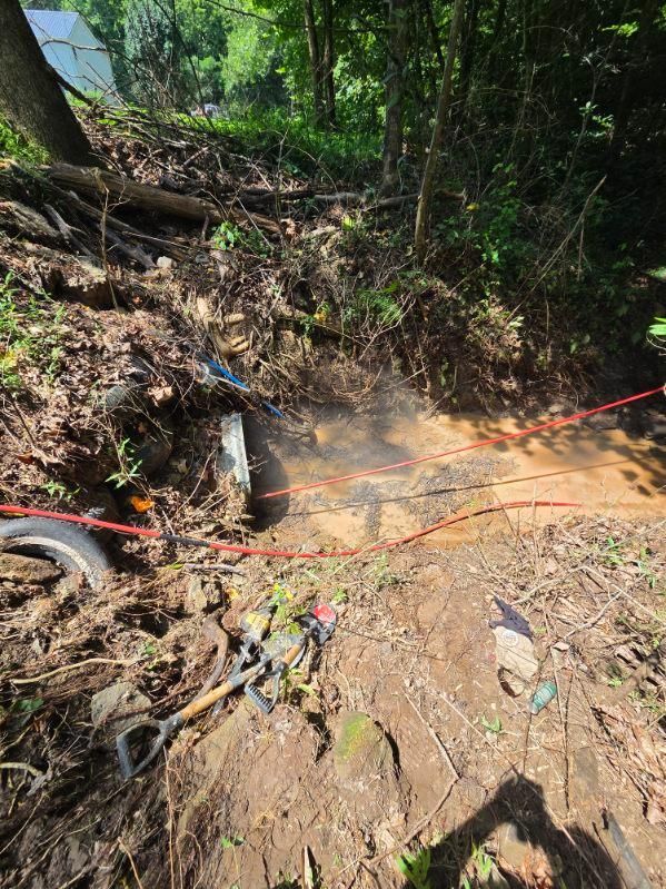 Steep muddy slope with a small water flow, roots, and red caution tape in a wooded area