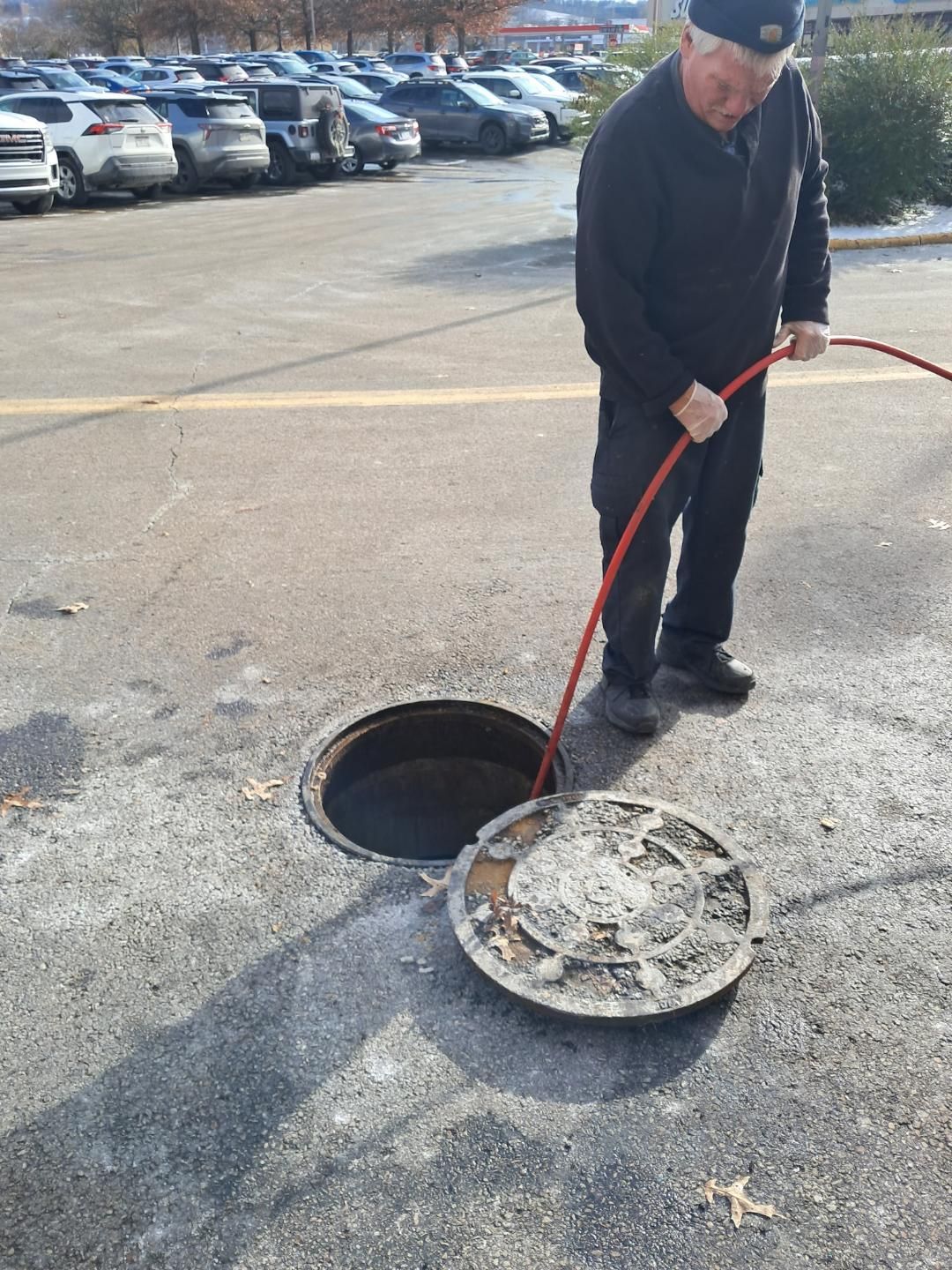 Worker cleaning a street manhole with a red hose in a parking lot