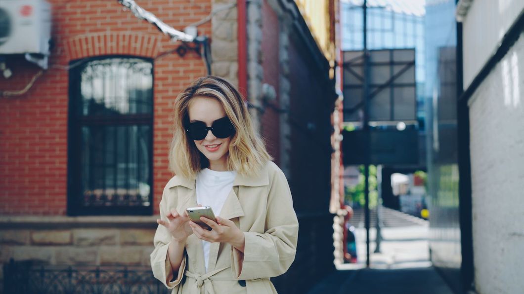 Woman in beige coat and sunglasses looking at her phone on a brick-lined city street