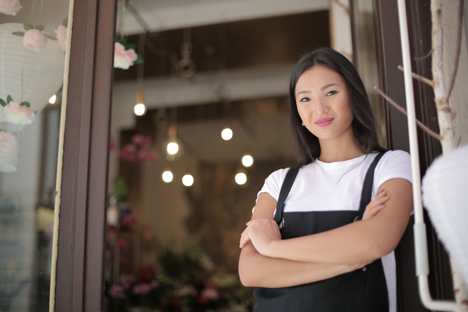 Person standing with arms crossed in a doorway, wearing a white shirt and black apron, with blurred lights behind them