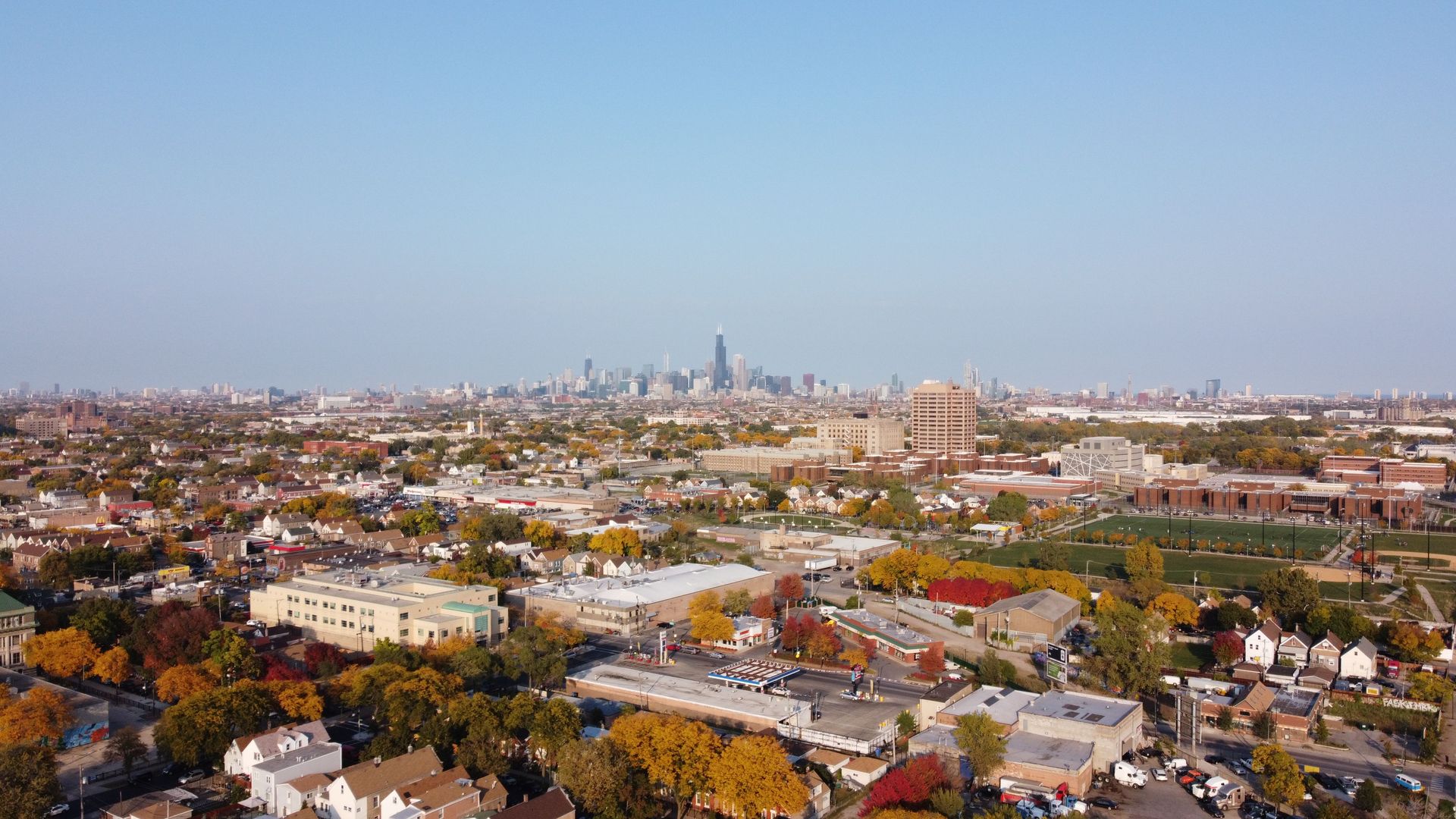 Aerial view of a suburban cityscape with trees, houses, and distant downtown skyscrapers under a clear sky