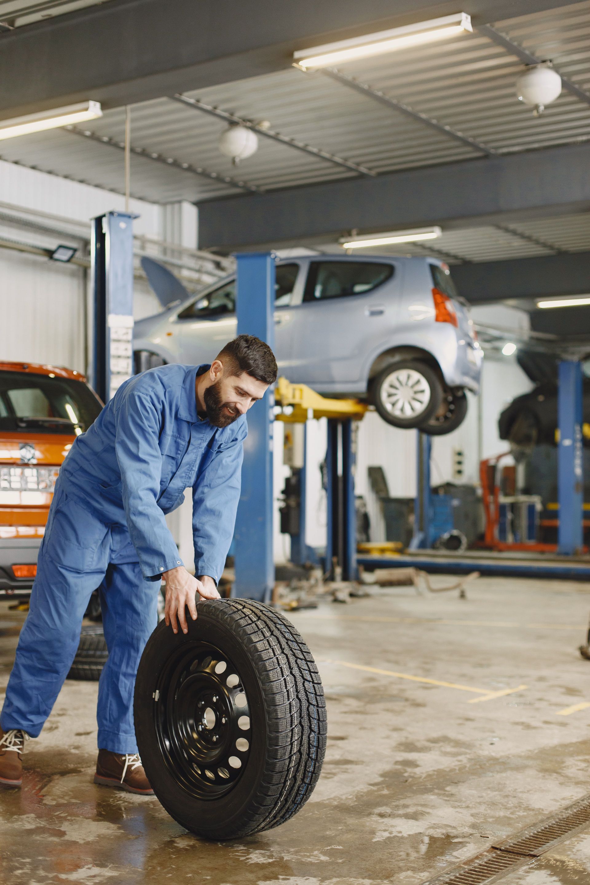 Mechanic in blue coveralls handling a tire in an auto repair shop with a car lifted on a hoist