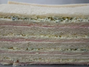 Sándwich de capas con jamón y lo que parece ser queso o relleno de ensalada de huevo entre rebanadas de pan.