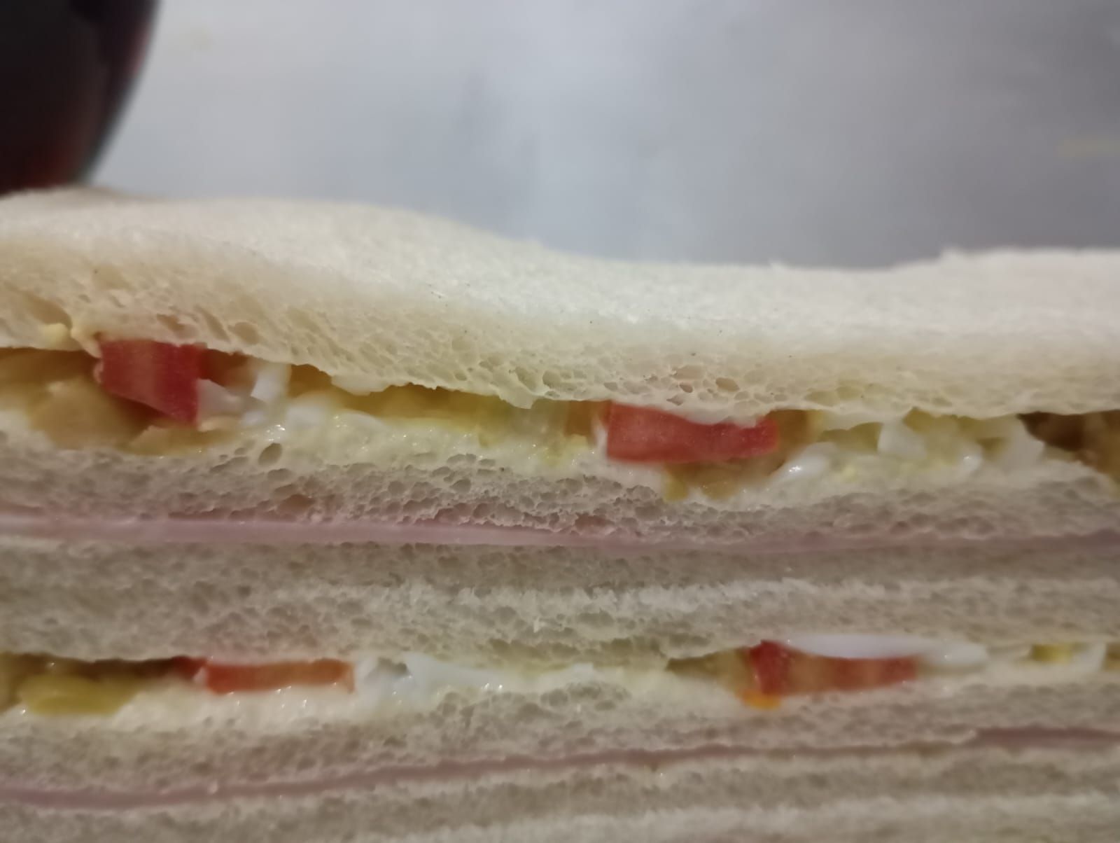 Primer plano de un sándwich con dos capas de jamón, tomates y una crema untable entre pan blanco.