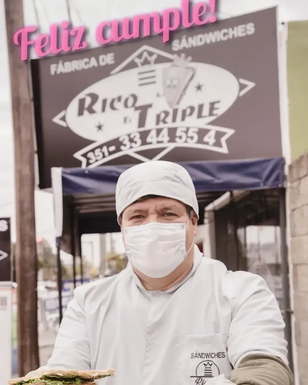 Hombre con bata de chef blanca y mascarilla sosteniendo un sándwich frente a un cartel de una tienda que dice