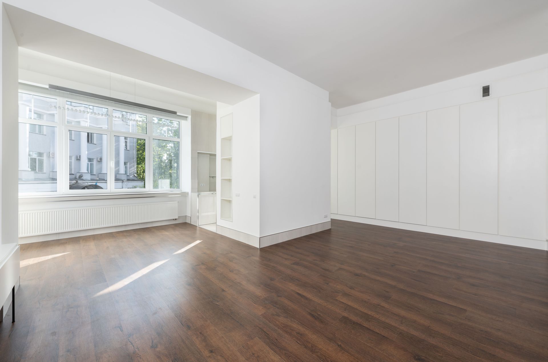 Empty white room with dark wood floors, large bay window, and built-in storage shelves.