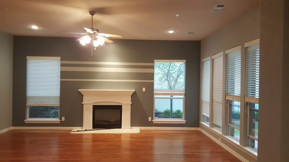 Empty living room with hardwood floors, gray walls, a fireplace, and large windows with blinds.