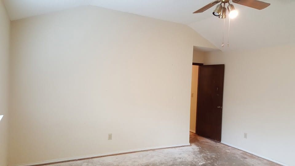 Empty beige bedroom with white walls, carpet, ceiling fan, and open doorway to a hallway