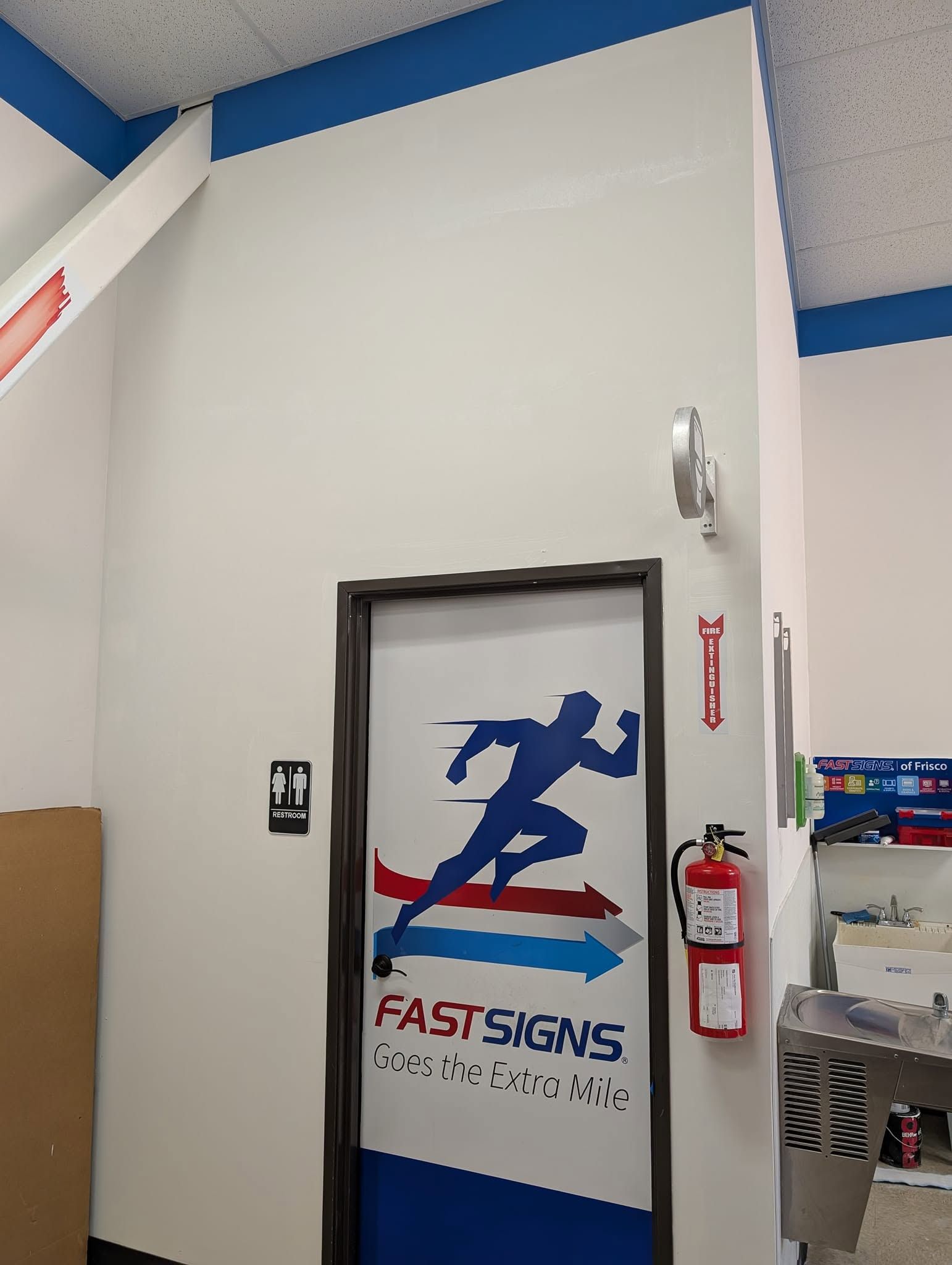 Hallway door with FastSigns logo and a fire extinguisher mounted beside it.
