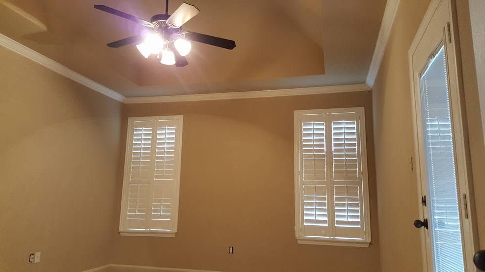 Empty beige room with tray ceiling, ceiling fan, two shuttered windows, and a glass door.