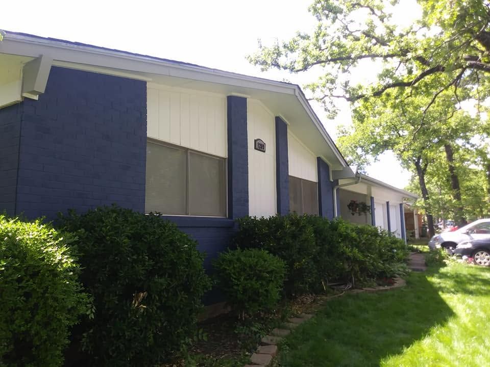 Blue apartment building with bushes and a grassy lawn beside a parked car under trees