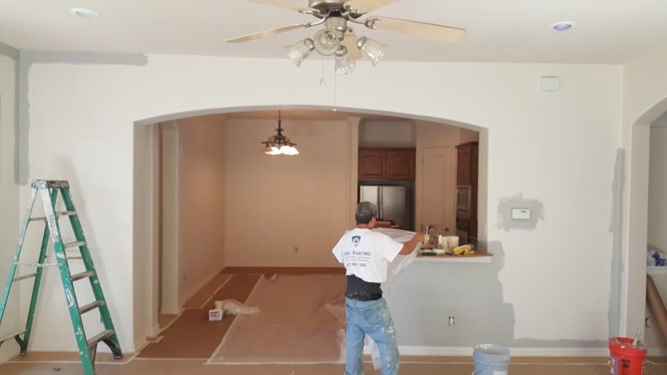 Home interior under renovation with a worker, ladder, and open arched pass-through to a kitchen.