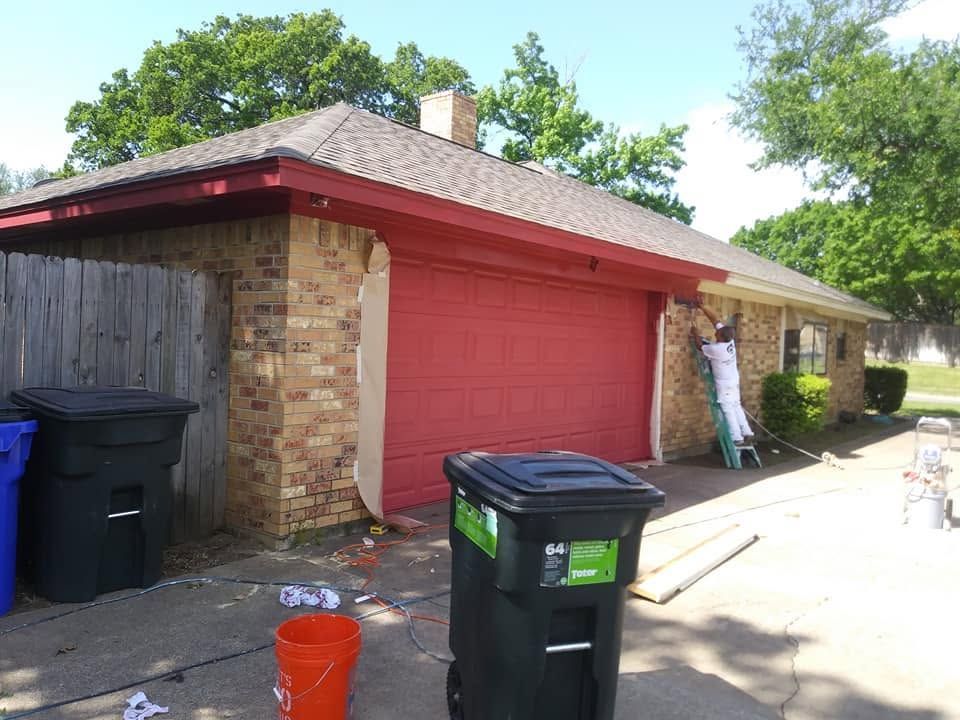 Brick garage with a red door, trash bins, and a ladder beside the house in a driveway