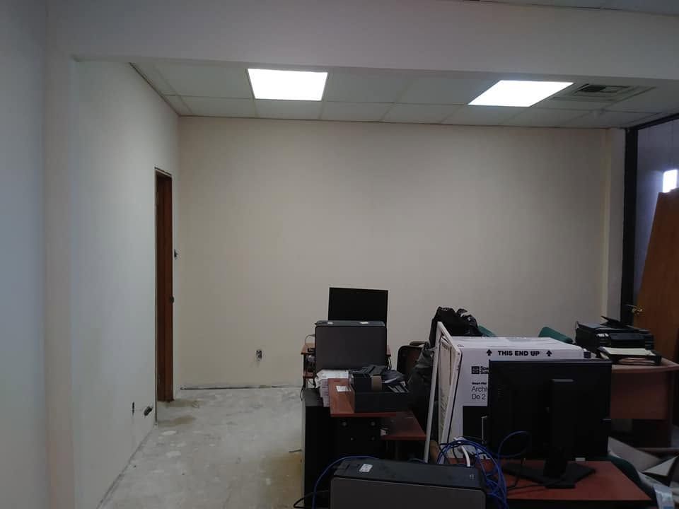 Empty office with white walls, fluorescent lights, and desks with computer equipment along the right side.